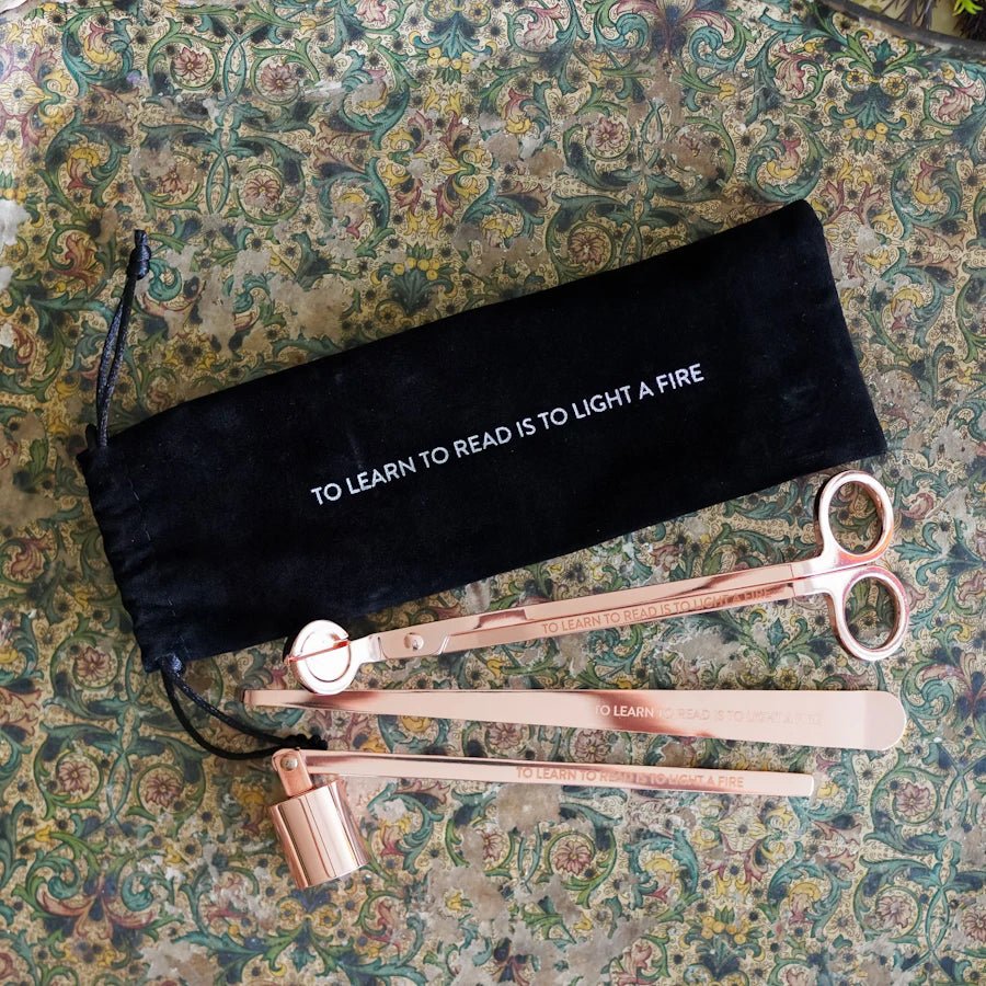 Bookish Candle Care Kit in velvet bag; rose gold snuffer, wick trimmer, dipper; reads: "TO LEARN TO READ IS TO LIGHT A FIRE."