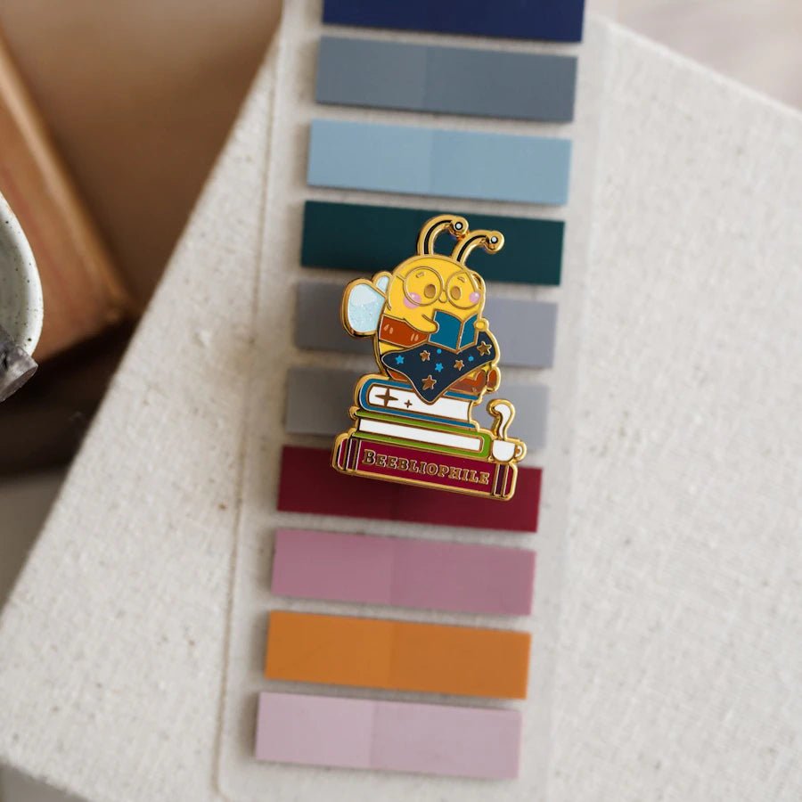 Beebliophile Enamel Pin has a yellow bee sitting on a stack of books while reading. One book is titled Beebliophile.