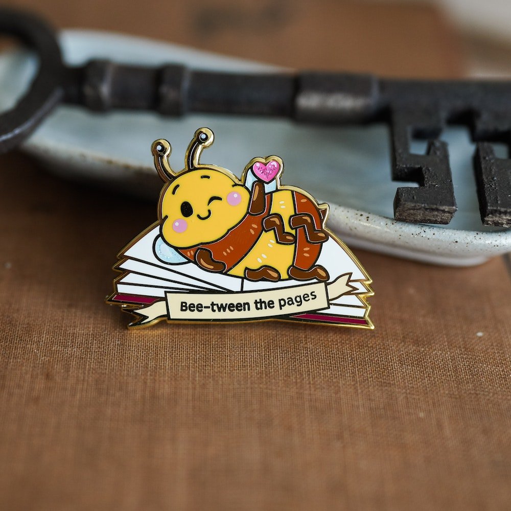 Book Bee Blind Bag Enamel Pin with images of a bee with text saying &quot;Bee-tween the pages&quot;