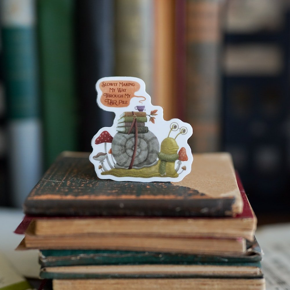 TBR Snail Sticker with books and a teacup strapped to its shell, mushrooms around the snail, and a quote bubble