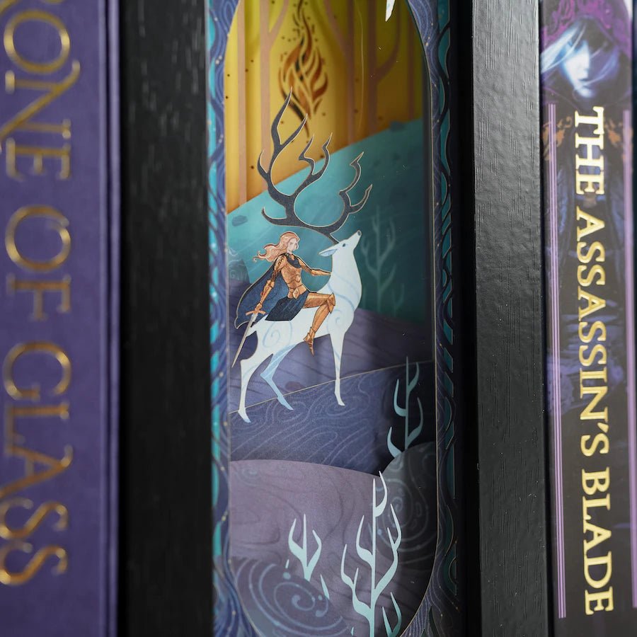 Purple, blue, and yellow Throne of Glass Bookshelf Alley is a black-framed shadowbox with a white papercut design of a woman on a stag.