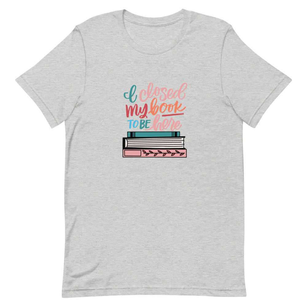 I Closed My Book To Be Here Short Sleeve Tee is a grey heather t-shirt with a colorful stack of books