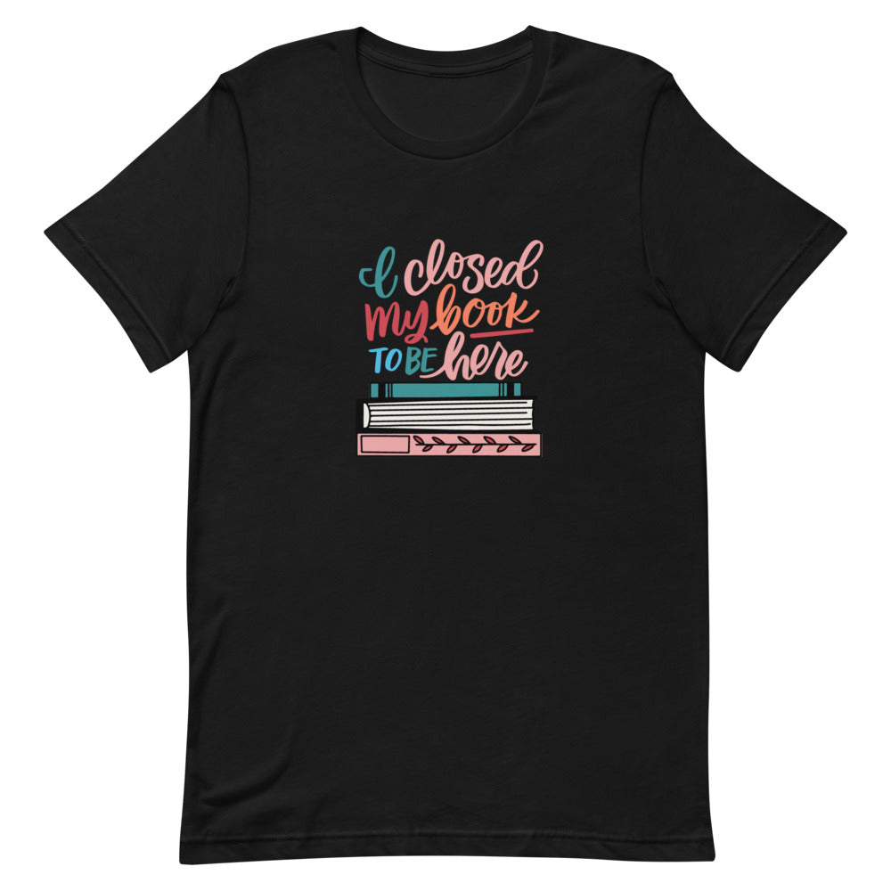I Closed My Book To Be Here Short Sleeve Tee is a black t-shirt with a colorful stack of books