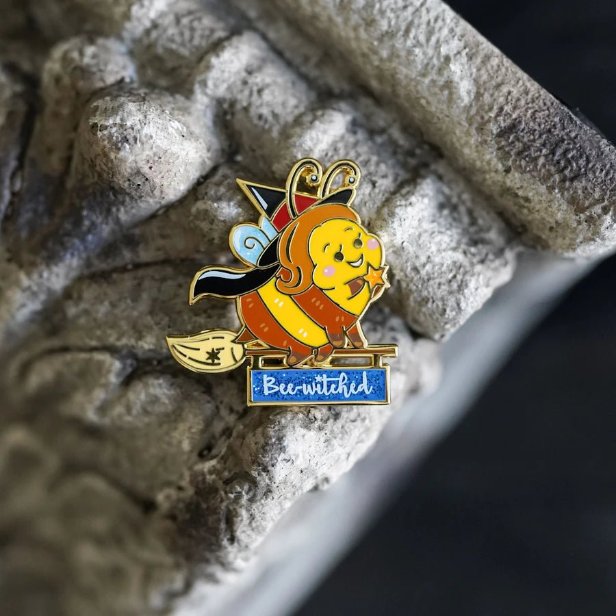 Bee-Witched Enamel Pin has a cute little bee on a broomstick with a witches hat, cloak, and a sign that reads "Bee-witched"