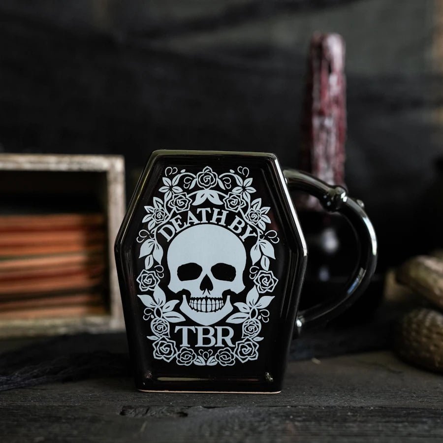 Death by TBR Mug is a black, coffin-shaped mug with a line drawing of a skull, flowers, and Death by TBR printed on the front.