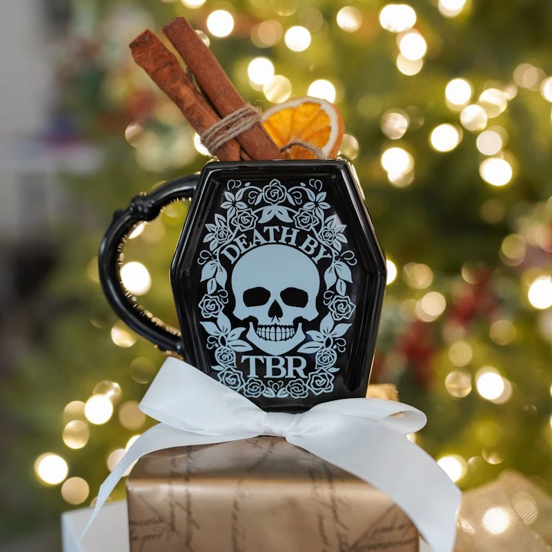 Death by TBR Mug is a black, coffin-shaped mug with a line drawing of a skull, flowers, and Death by TBR printed on the front.