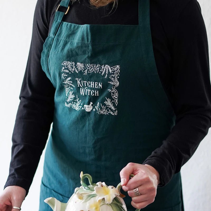 Kitchen Witch Apron is dark green with a white screen printed design featuring a pestle and mortar, herbs, and plants