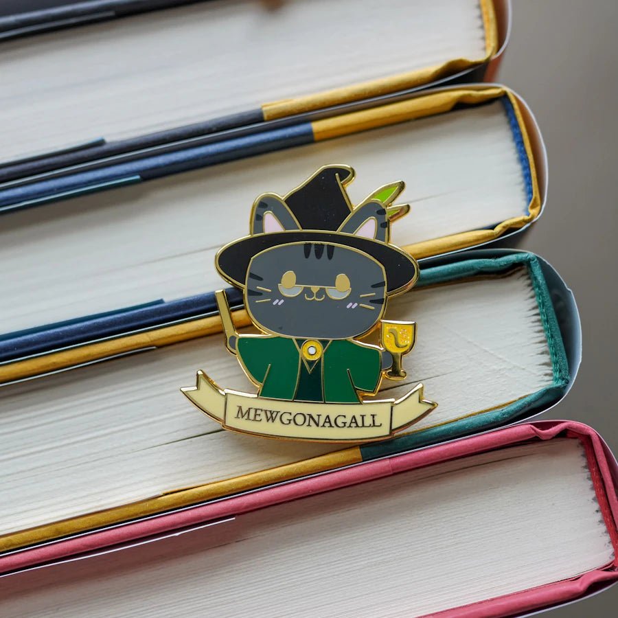 Mewgonagall Cat Enamel Pin is a grey cat dressed in a green wizard cloak, wearing a witch hat, and holding a wand and a goblet.