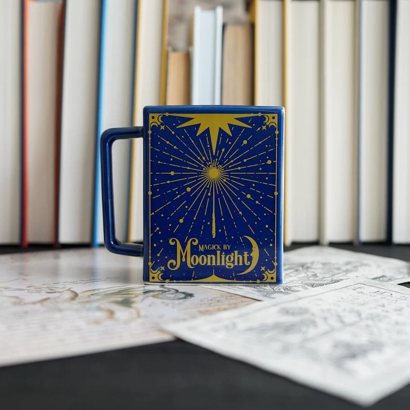 Navy Moon Magic Book Mug is shaped like a book with a handle and has golden yellow design with "Magick by Moonlight" on the shiny glaze.