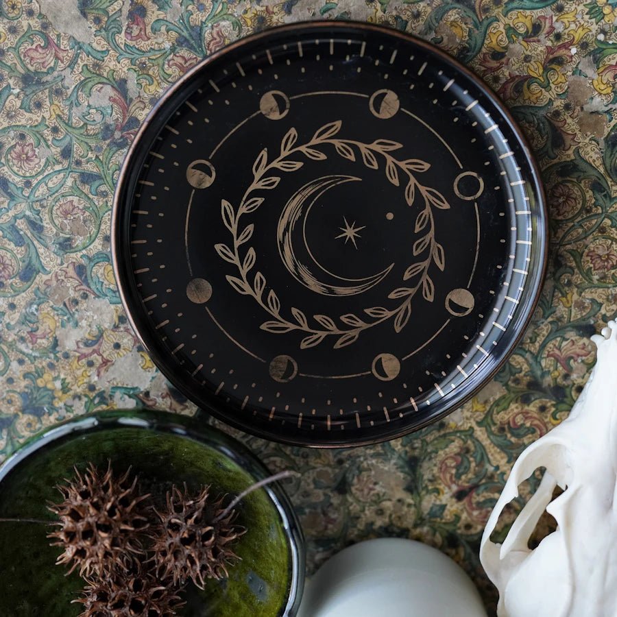 Moon Magic Trinket Dish—a black circular ceramic dish with metallic gold printing of moon phases and more celestial designs