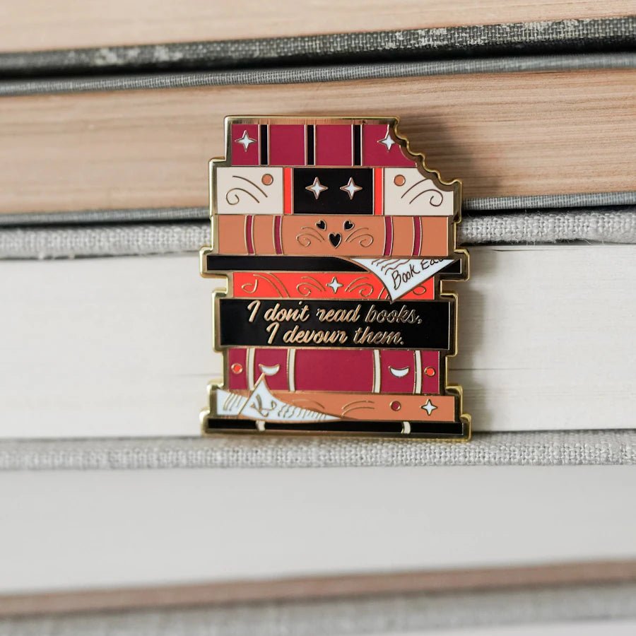 The Book Eaters enamel pin features a stack of books with a bite taken out—spine reads, &quot;I don&#39;t read books, I devour them.&quot;