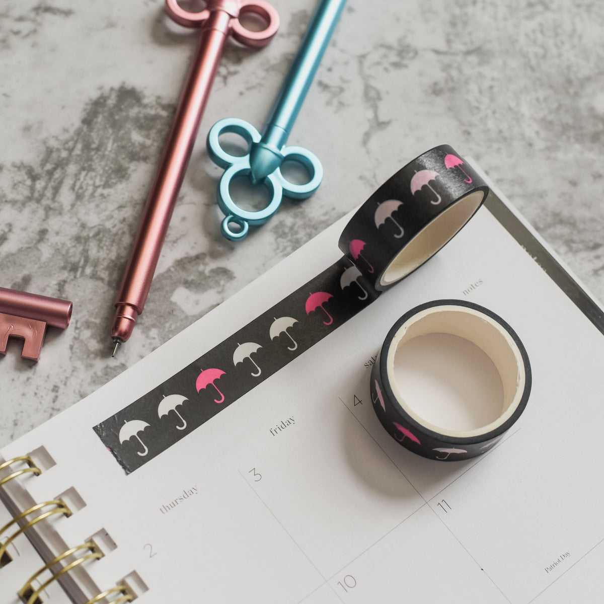 Umbrella Academy Washi Tape is a black roll of washi tape with gray, white, and pink umbrellas