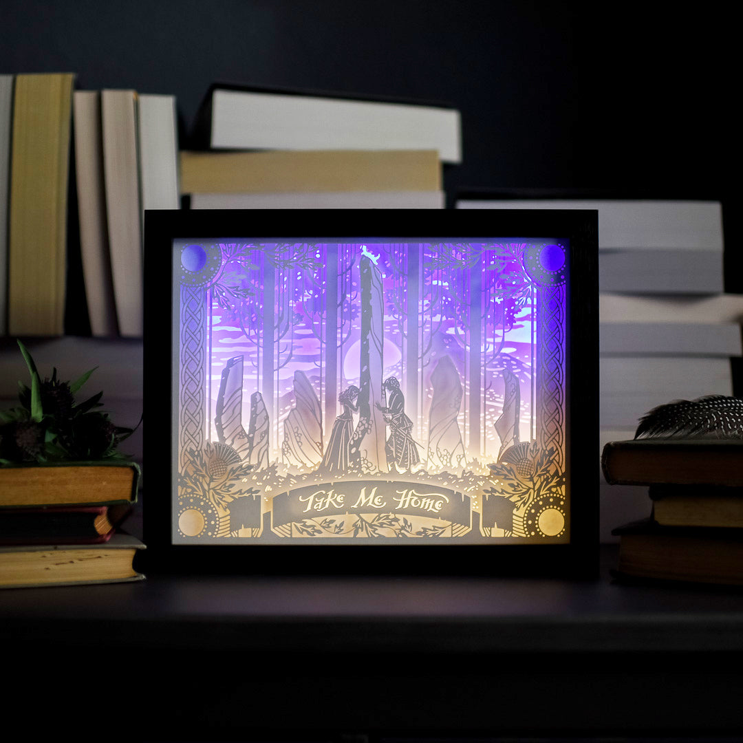Standing Stones Light Box is a black framed paper cut light box with purple and yellow light behind it and the words &quot;Take Me Home&quot;