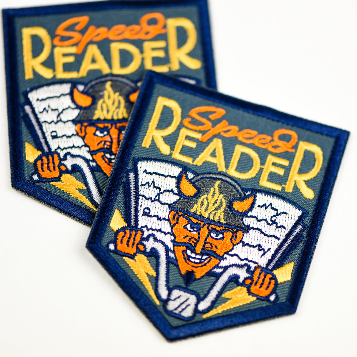 Speed Reader Patch is dark blue with orange and yellow &quot;Speed Reader&quot; and a orange faced devil on a bike with a book behind him