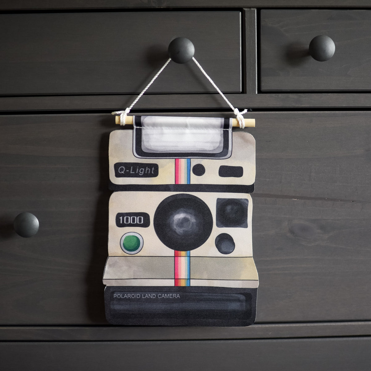 Polaroid Camera Pin Banner is a vintage off-white camera with a rainbow stripe down the middle made of fabric and hanging on a wooden dowel with a string