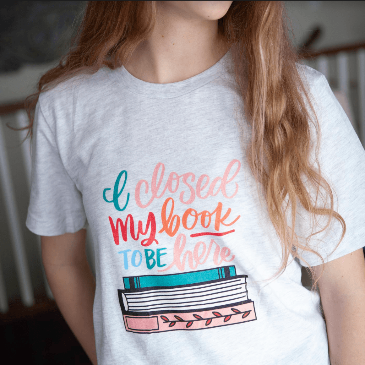 I Closed My Book To Be Here Short Sleeve Tee is a t-shirt with a colorful stack of books and the quote &quot;I closed my book to be here&quot;