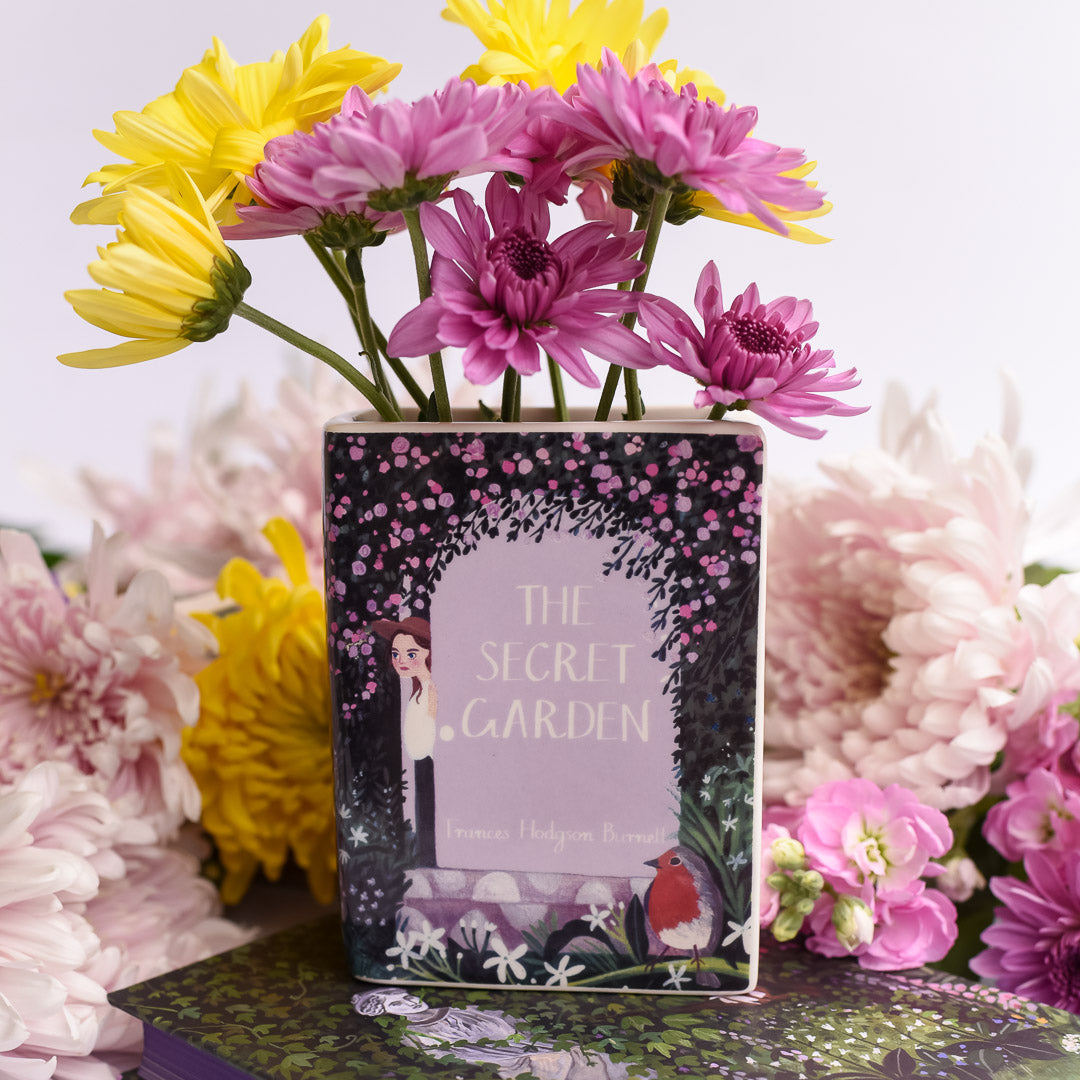 The Secret Garden Book Vase has artwork of a young girl peeking from behind a purple door with pink and purple flowers and garden aesthetic