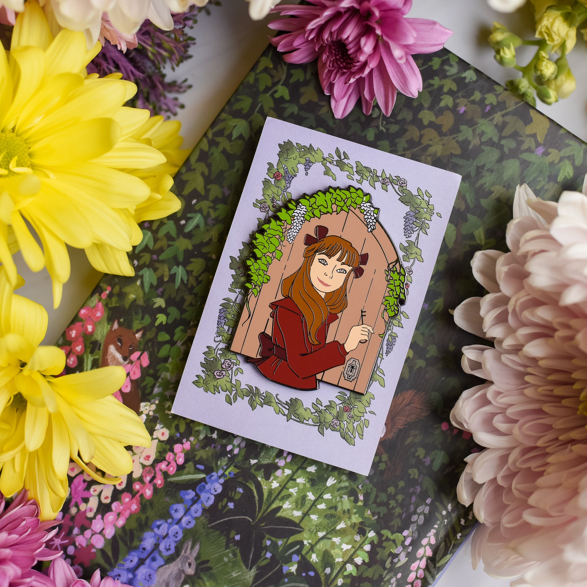 The Secret Garden Enamel Pin has Mary with a key standing in front of a door and surrounded by garden flowers