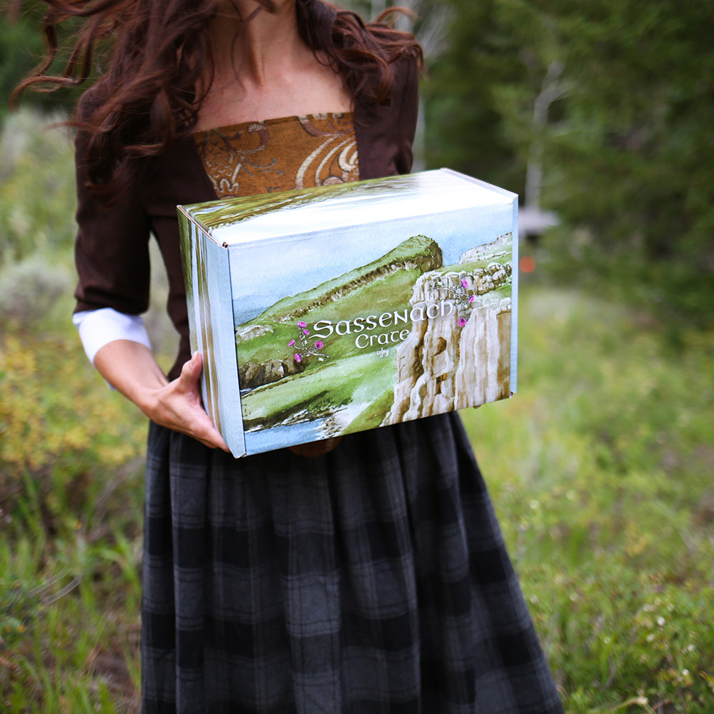 Sassenach Crate held by a woman that looks like Claire while standing on a grassy hill