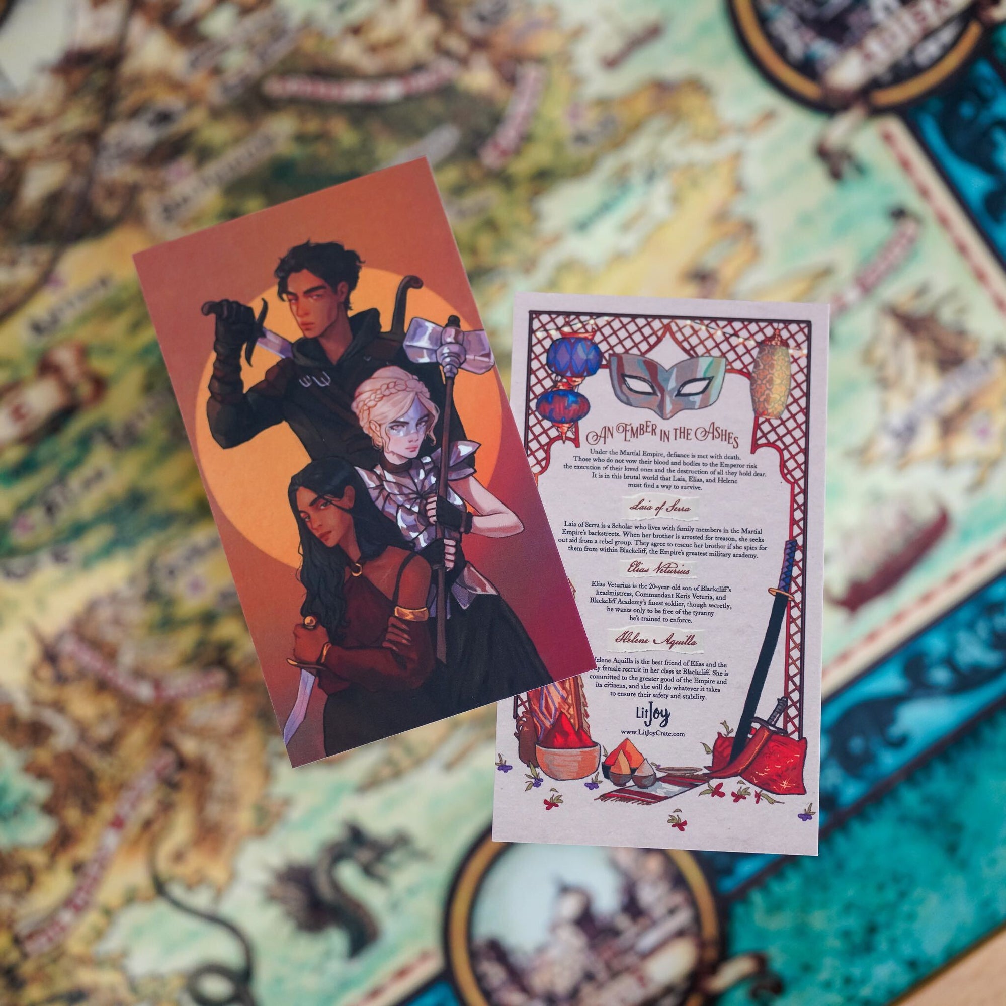 An Ember in the Ashes Adventure Card features Laia, Elias, and Helene holding weapons with character details on the back.