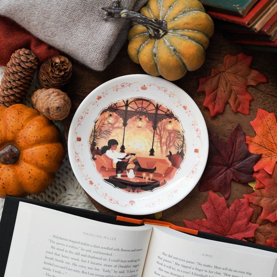 Autumn Readers Dessert Plate has autumn designs of pumpkins, leaves, a girl lounging on a couch reading, and a coffee table with tea, cookies, and candles.