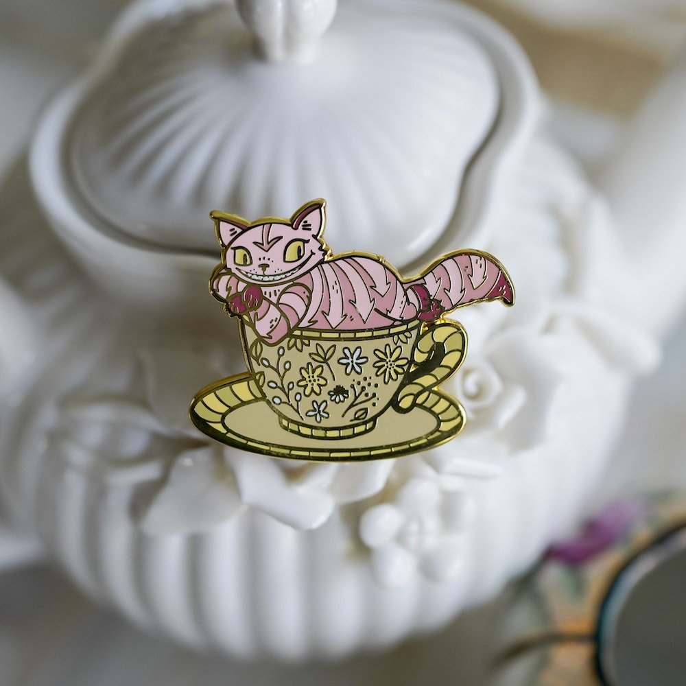 Chesire Cat Critter Collection Enamel Pin with a pink, striped, smiling cat sitting in a teacup on top of a saucer.