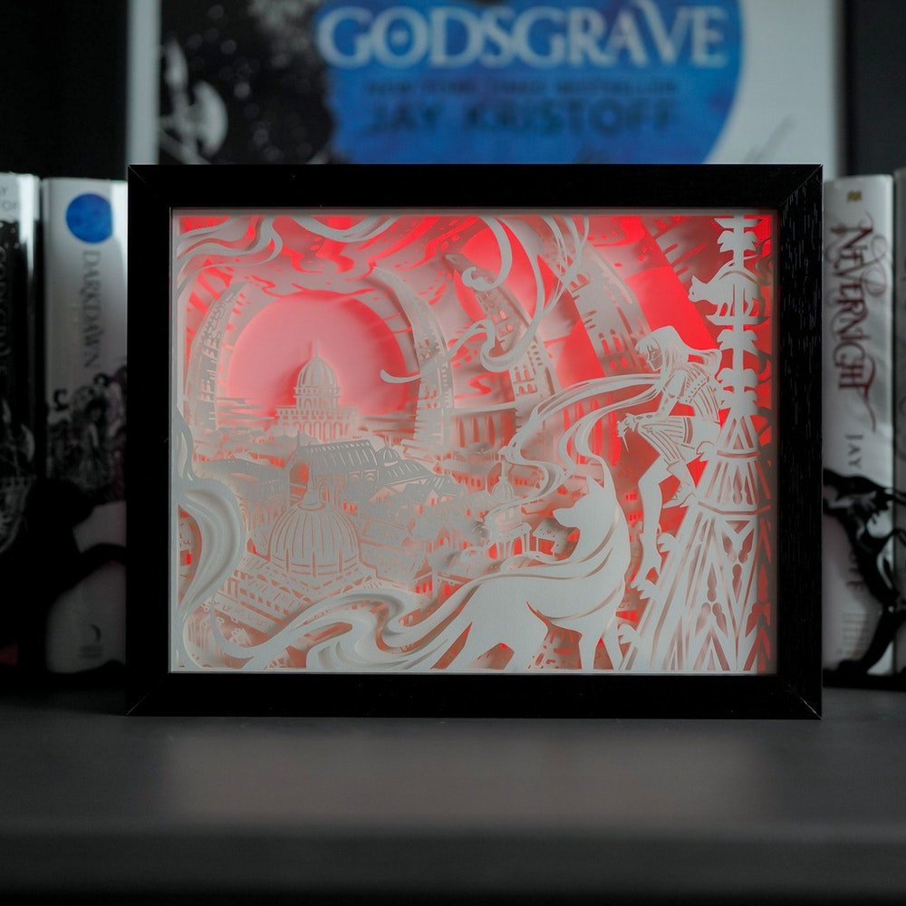 Darkin Shadow Light Box shows a paper cut shadowbox with red light of Mia Corvere and shadow daemons overlooking Godsgrave from Nevernight by Jay Kristoff