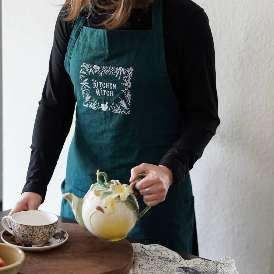 Kitchen Witch Apron is dark green with a white screen printed design featuring a pestle and mortar, herbs, and plants