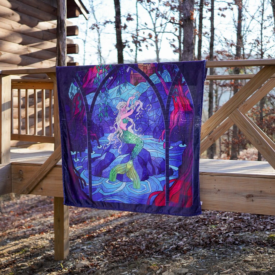 Stained-glass designed Mermaid Tapestry Blanket with a mermaid on a rock in water amid shades of blue and red background