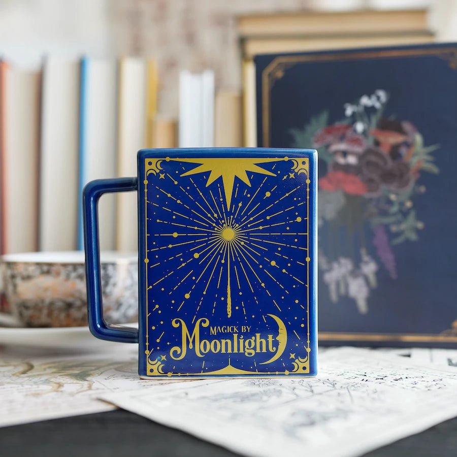 Navy Moon Magic Book Mug is shaped like a book with a handle and has golden yellow design with &quot;Magick by Moonlight&quot; on the shiny glaze.