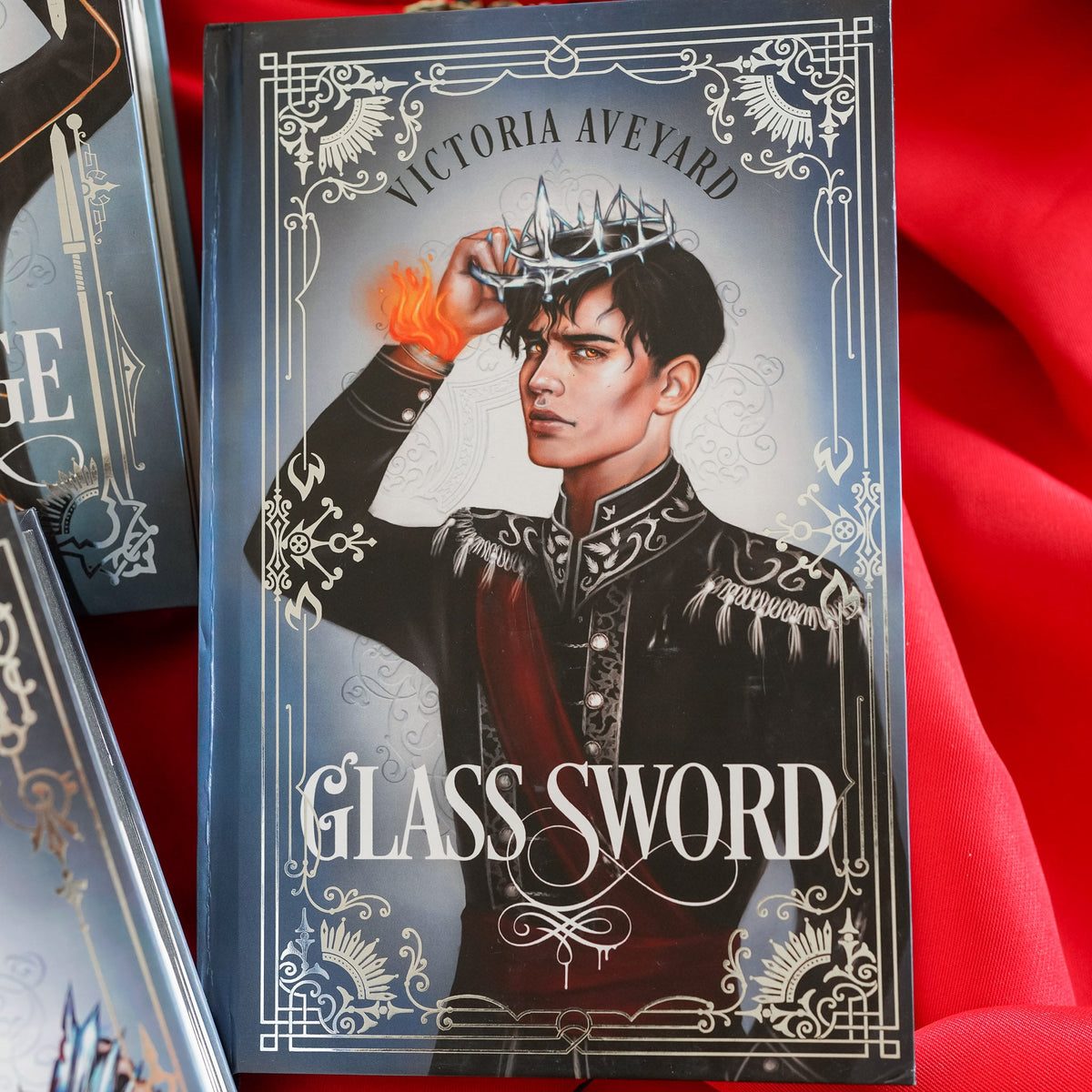 Glass Sword book 2 featuring Tiberias Calore VII with crown in his hand and flames coming from his fire-starter bracelet