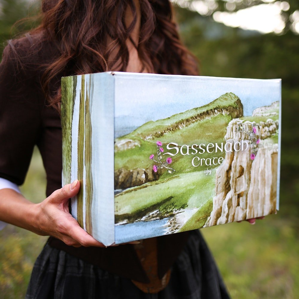 Sassenach Crate held by a woman that looks like Claire while standing on a grassy hill