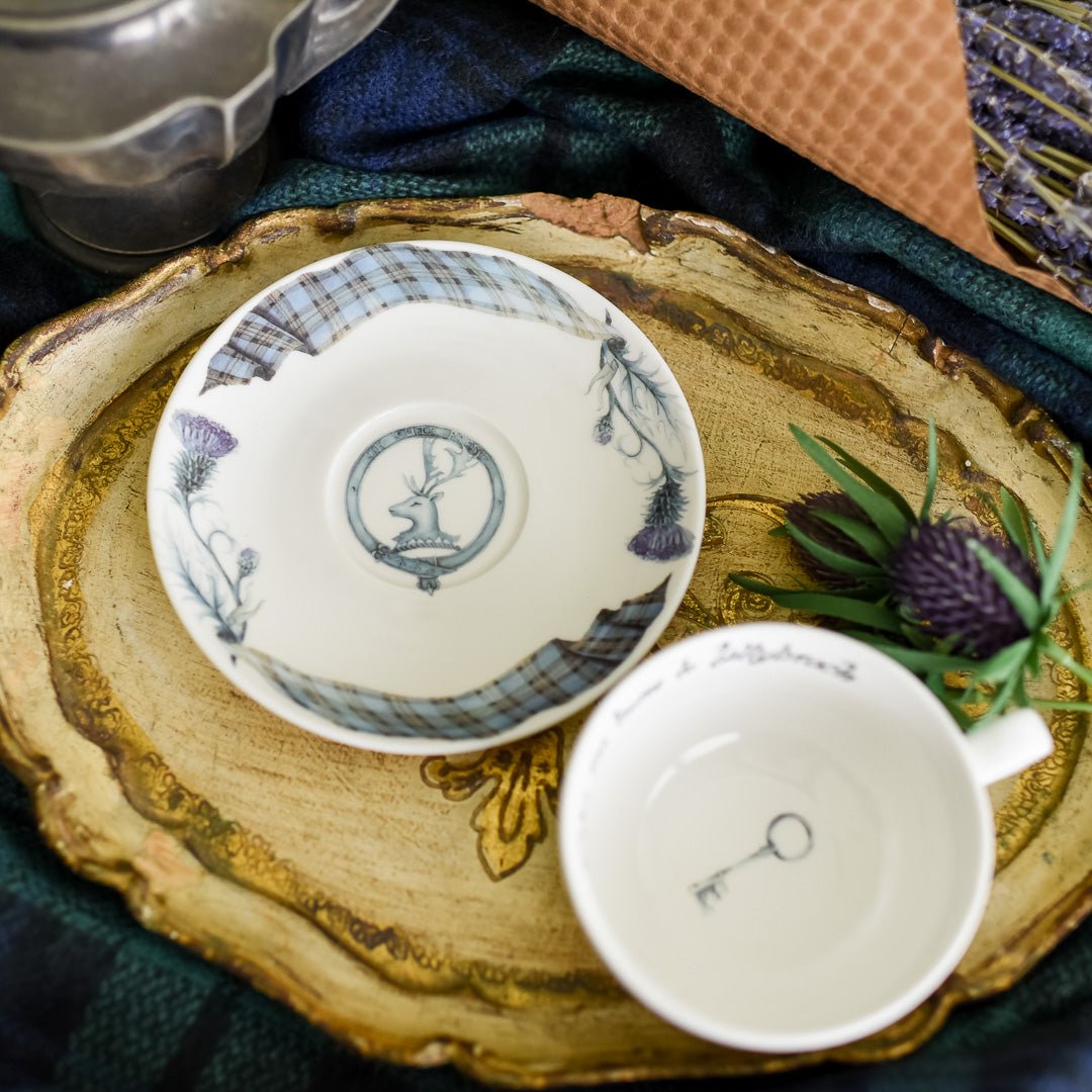 A white Sassenach Teacup and Saucer with tartan pattern, blue flowers, and Scottish family crest with words Je suis prest