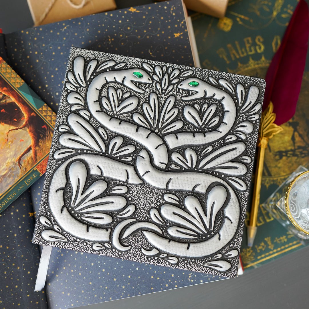 Snake Chamber Notebook is a square shaped notbook with a faux metal cover with two intertwined snakes with green gem eyes
