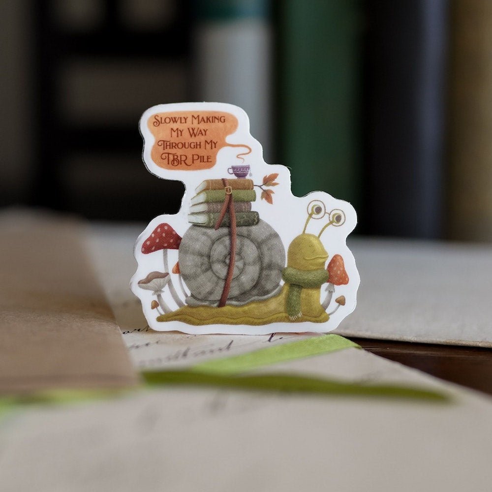 TBR Snail Sticker with books and a teacup strapped to its shell, mushrooms around the snail, and a quote bubble