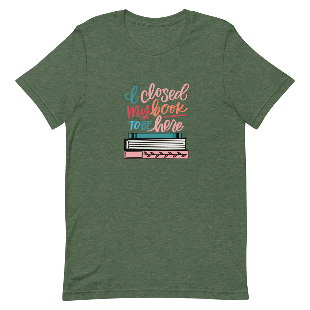 I Closed My Book To Be Here Short Sleeve Tee is a green t-shirt with a colorful stack of books