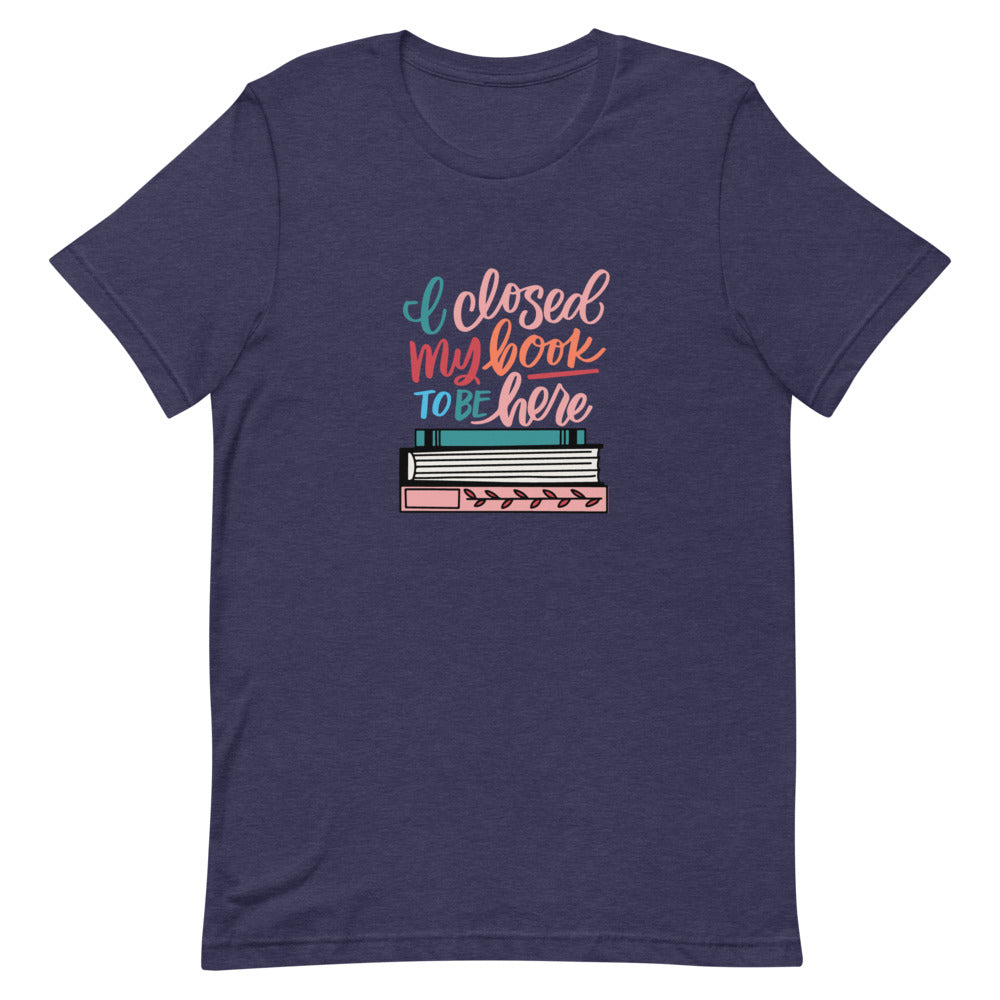 I Closed My Book To Be Here Short Sleeve Tee is a navy blue t-shirt with a colorful stack of books