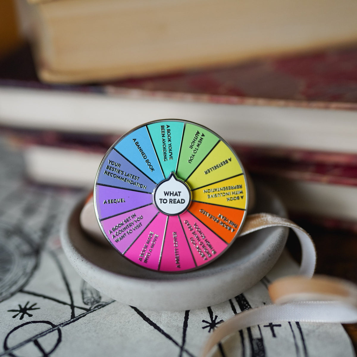 What To Read Spinner Enamel Pin with colored labels listing various book choices.