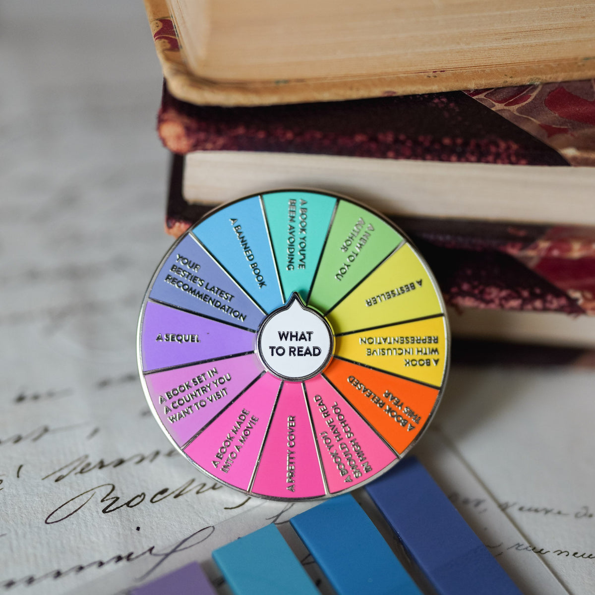 What To Read Spinner Enamel Pin with colored labels listing various book choices.